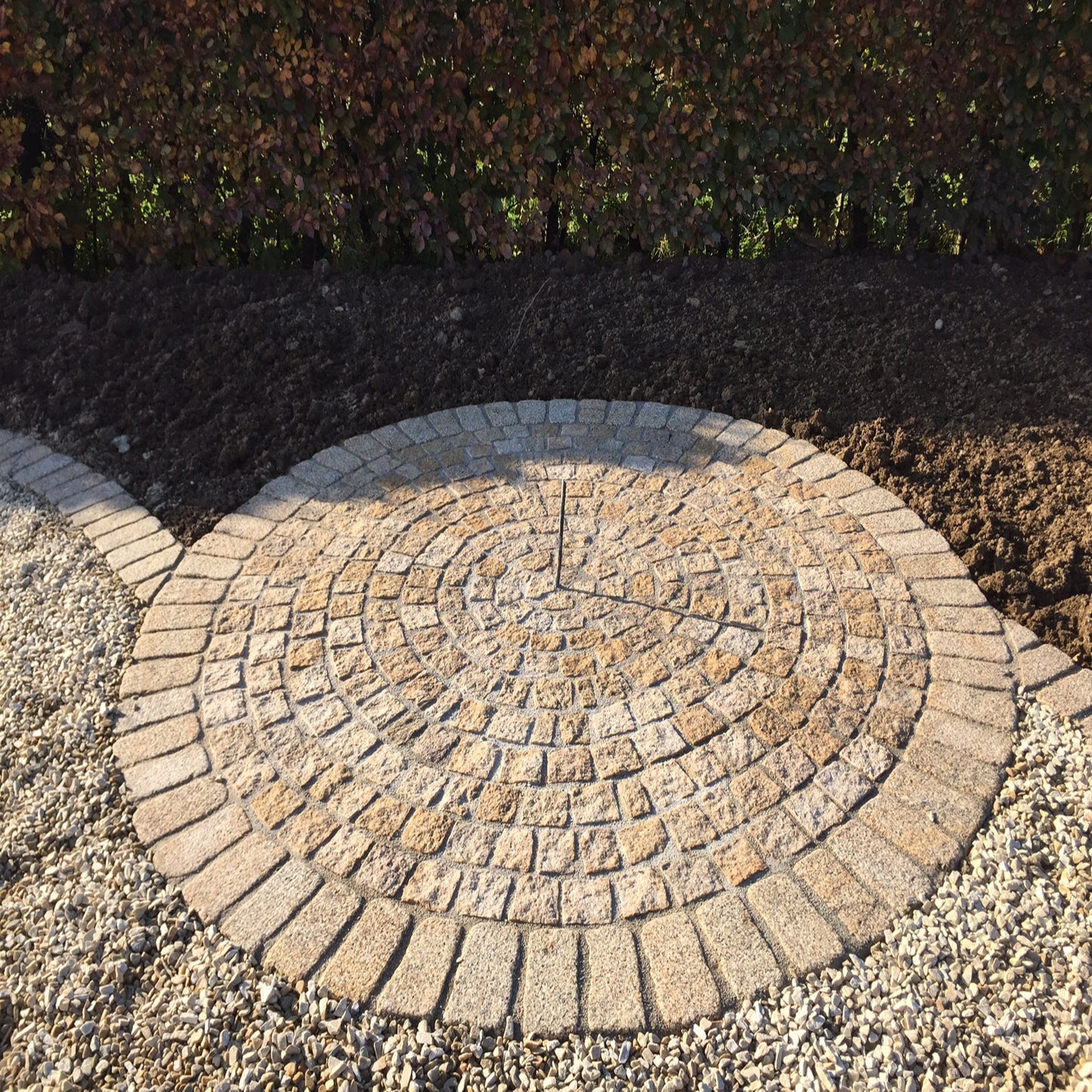 A circular pattern of gold granite paving cobbles with a bush hammered surface and lightly tumbled edges installed on a bed of gravel. Inner cobbles are Gold Granite Paving Cobbles in 100 x 100mm size. with outer circle in 200 x 100mm size.