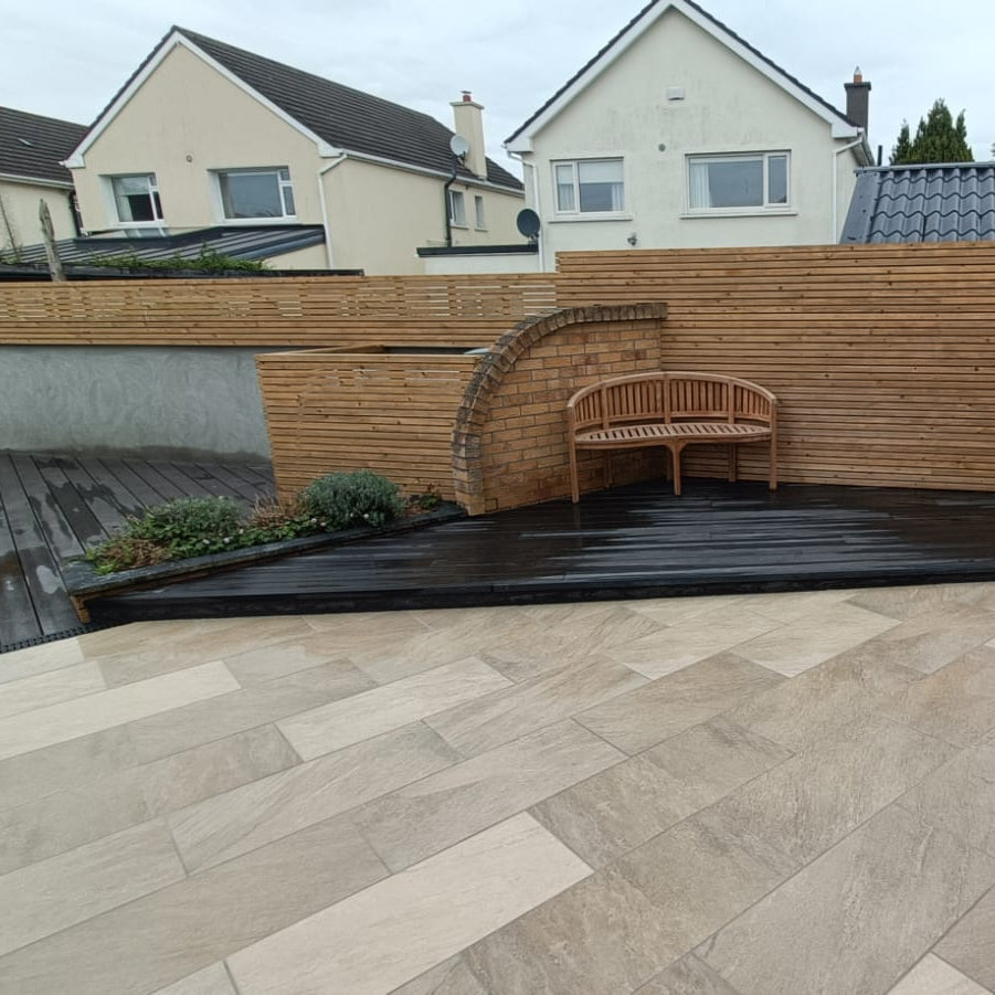 Garden with wooden bench, stone patio, and houses in the background. Patio completed using Gold Quartz Dive Range Porcelain Paving.