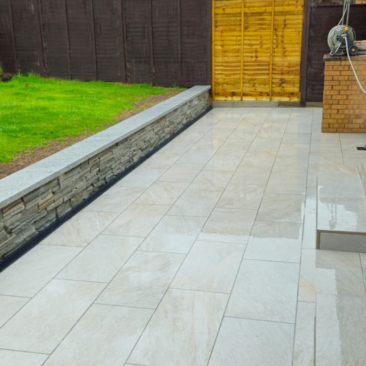 Paved patio area finished with Silver Quartz Diva Range Porcelain Paving with a stone wall and wooden fence in the background