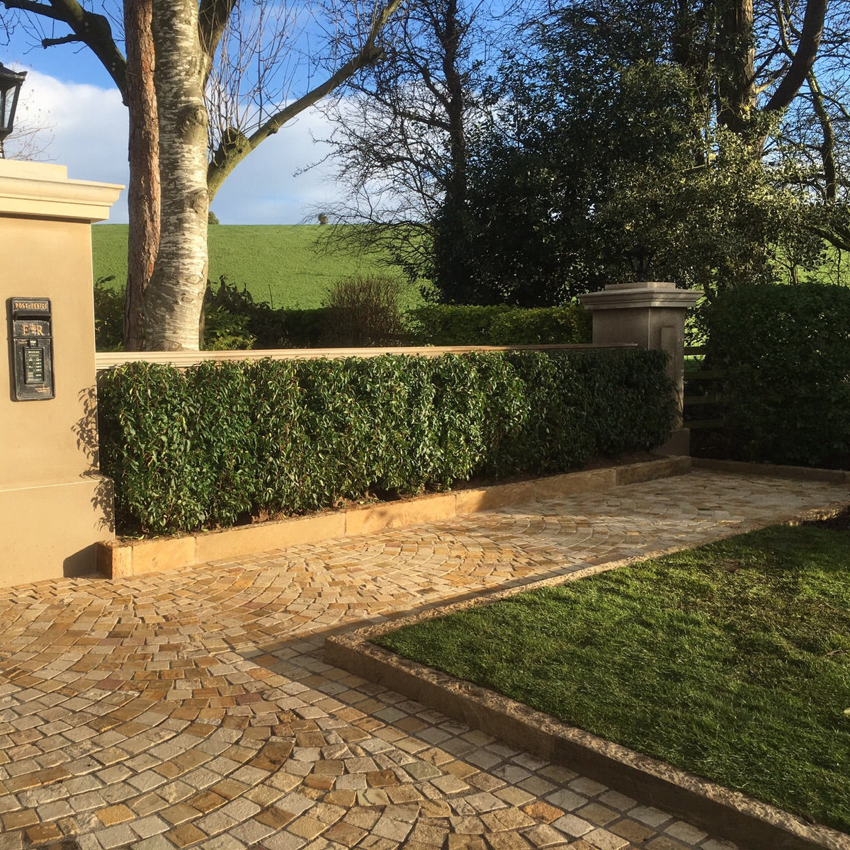Paved driveway leading to a gate with a hedge and tree in the background. Driveway and pathway edging completed using Tinted Mint Sandstone Kerbs.