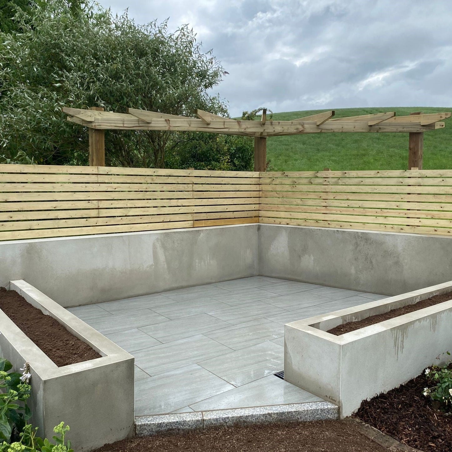 Small patio area with raised flower beds and wooden fence. Paving is Ashen Jewel Range Porcelain Paving Tiles in 900 x 600 x 20mm.