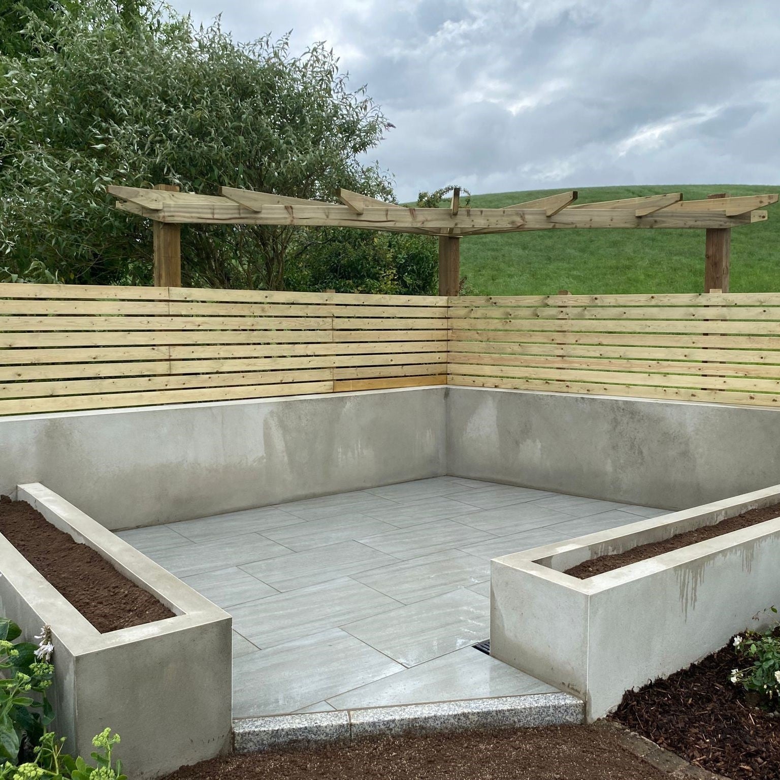 Small patio area with raised flower beds and wooden fence. Paving is Ashen Jewel Range Porcelain Paving Tiles in 900 x 600 x 20mm.