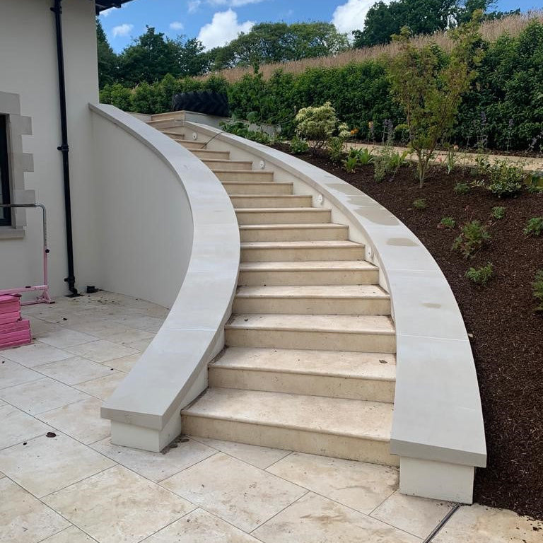 Curved bespoke sandstone stairs leading up to a house with a clear blue sky and greenery in the background.