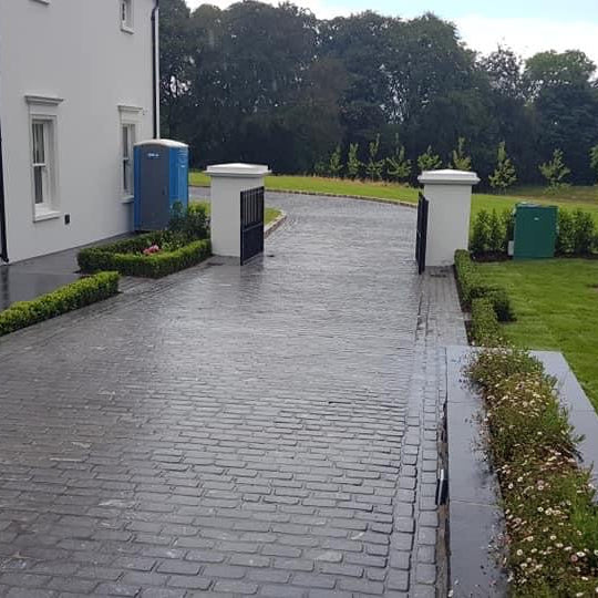 Paved driveway using Black Granite Cobbles leading to a house with greenery and trees in the background