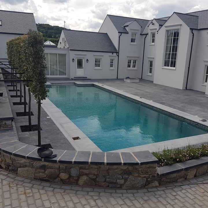 Swimming pool in front of a modern house with grey roofs and white walls. Driveway in the foreground is paved using Black Granite Cobbles in 200 x 100mm size.