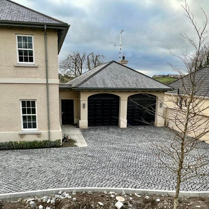 Two-story house with a driveway paved with Black Granite Cobbles, surrounded by trees and a cloudy sky.