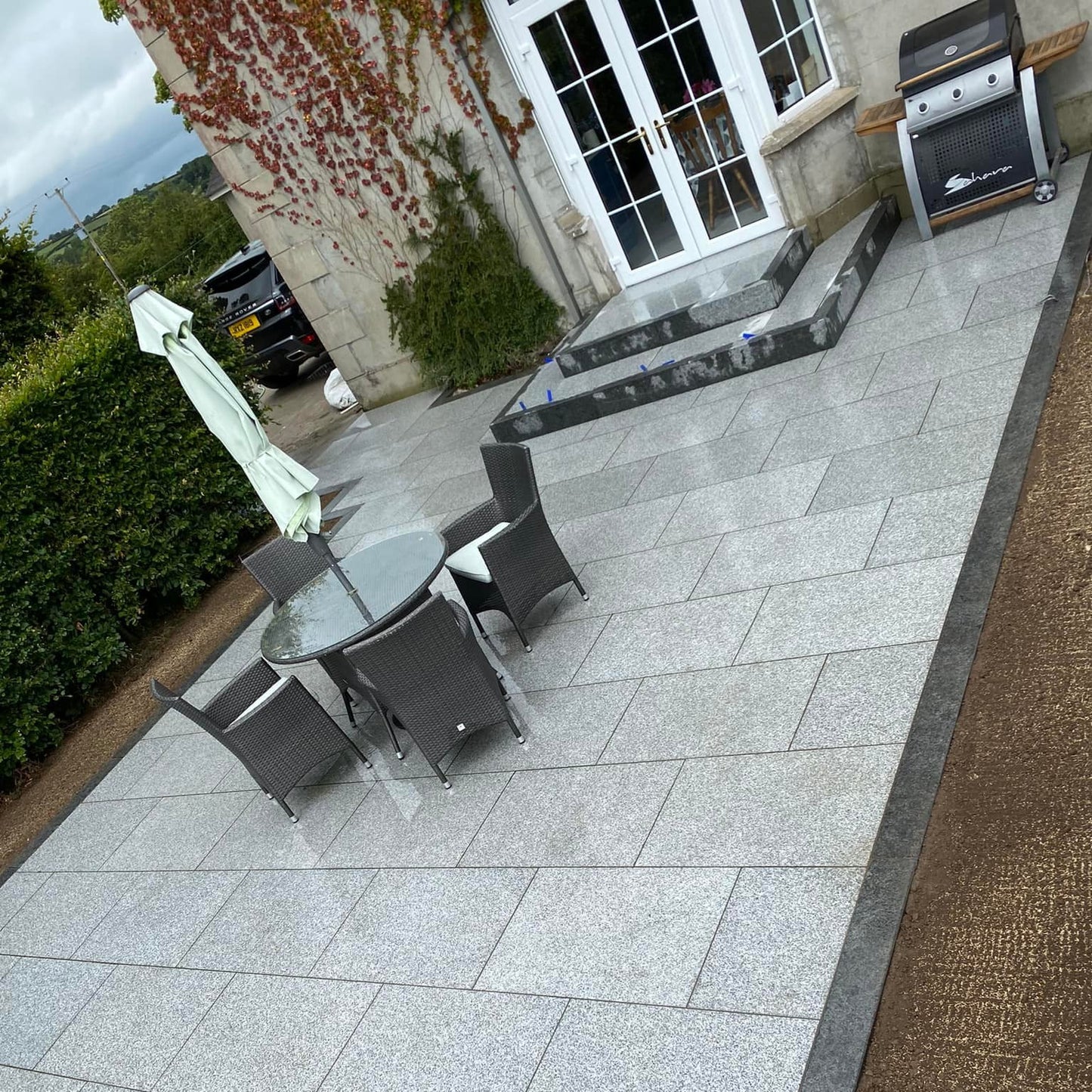 Outdoor patio area with a glass table and chairs on a stone patio. Patio steps and border completed using Black Granite Pin Kerbs. 