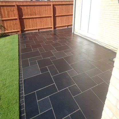 A completed patio installation using black limestone paving with a wooden fence and artificial grass in the background.