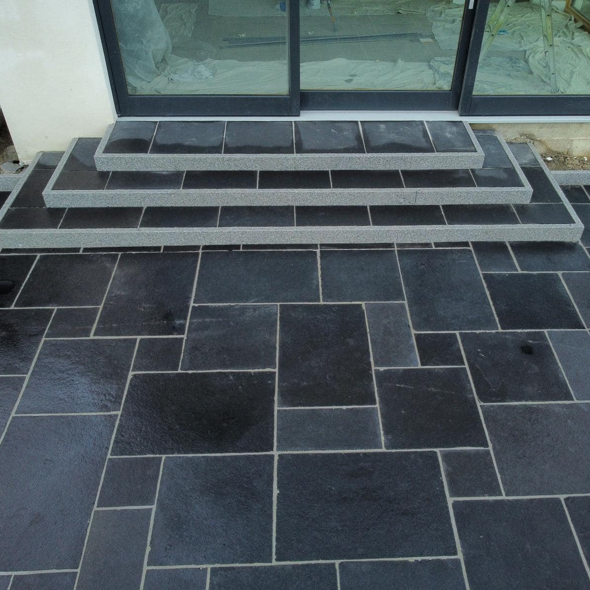 Black limestone patio slabs arranged on the ground in a pattern, with a set of steps leading up to patio doors. 