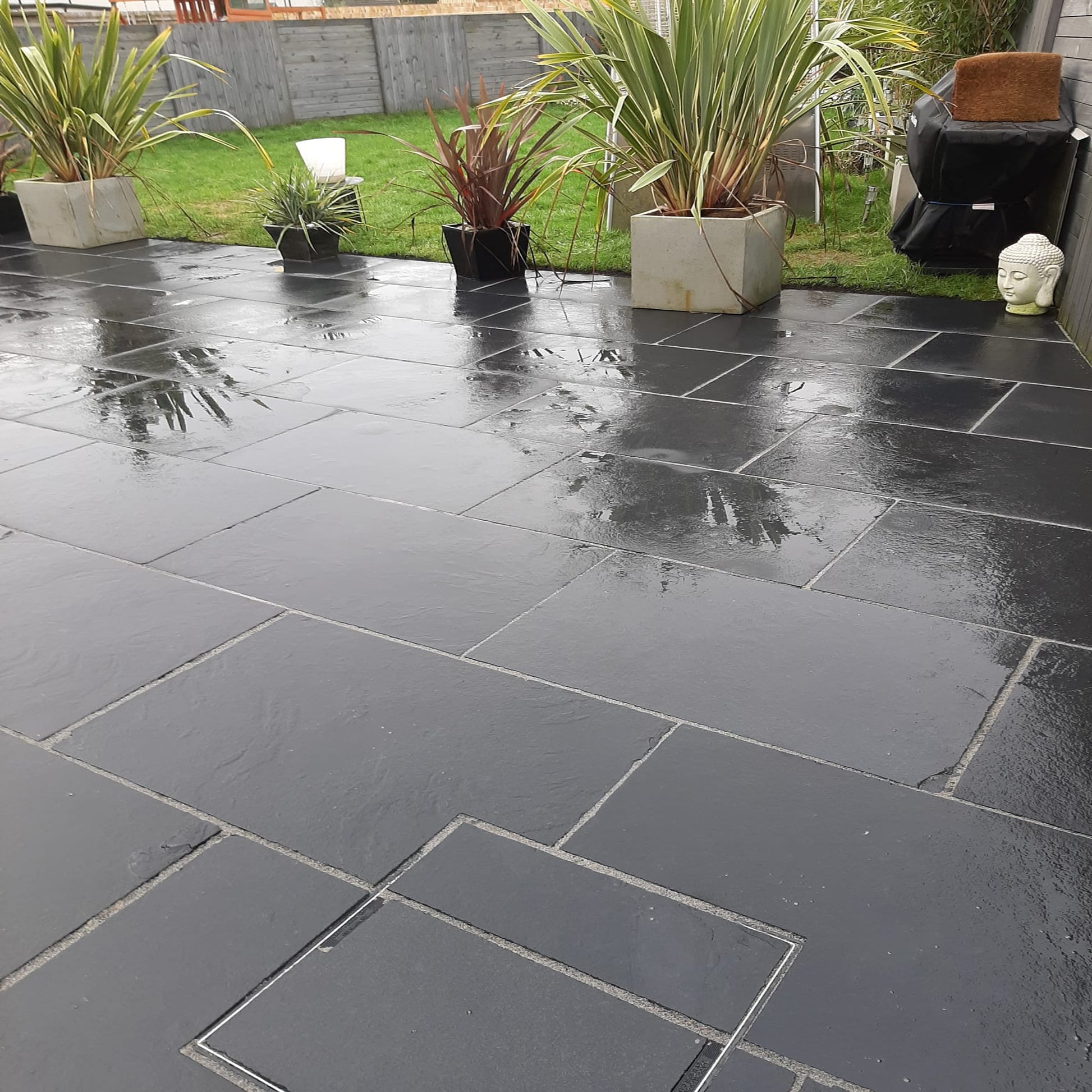 Black limestone paving slabs arranged on a patio with potted plants and outdoor furniture in the background.