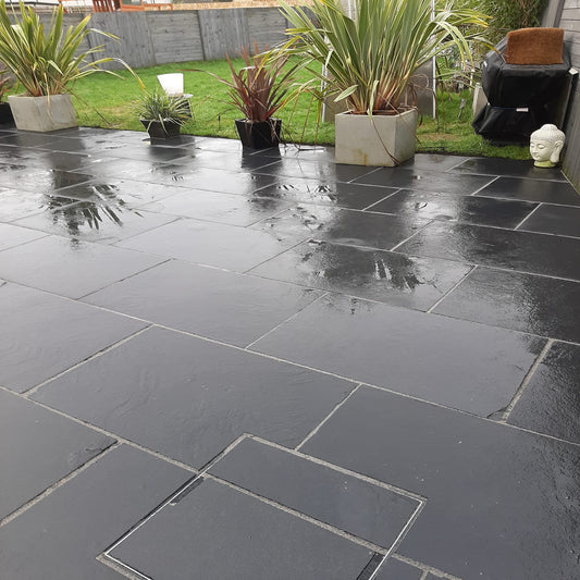 Black limestone paving slabs arranged on a patio with potted plants and outdoor furniture in the background.
