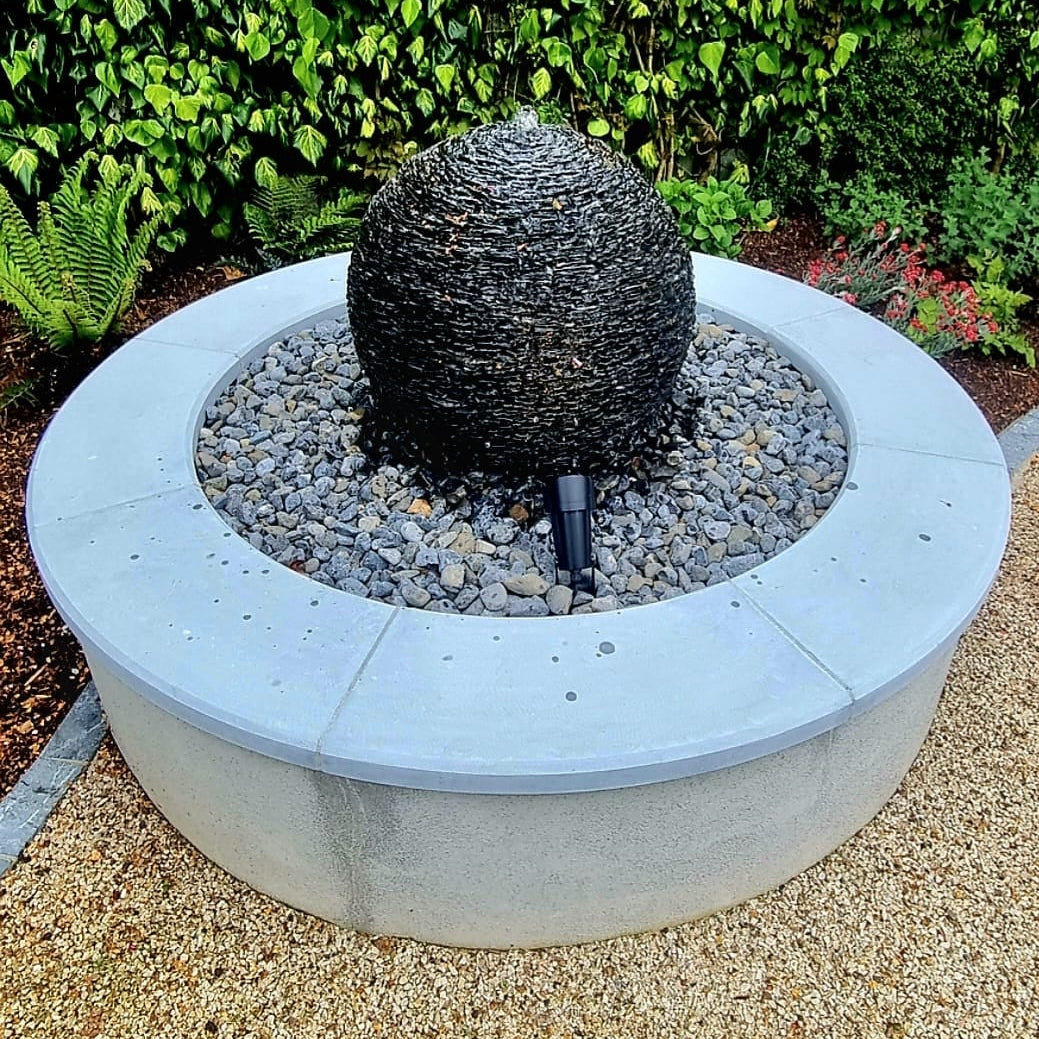 Decorative stone sphere in a circular stone planter with greenery in the background. Wall completed with curved blue limestone wall capping. 