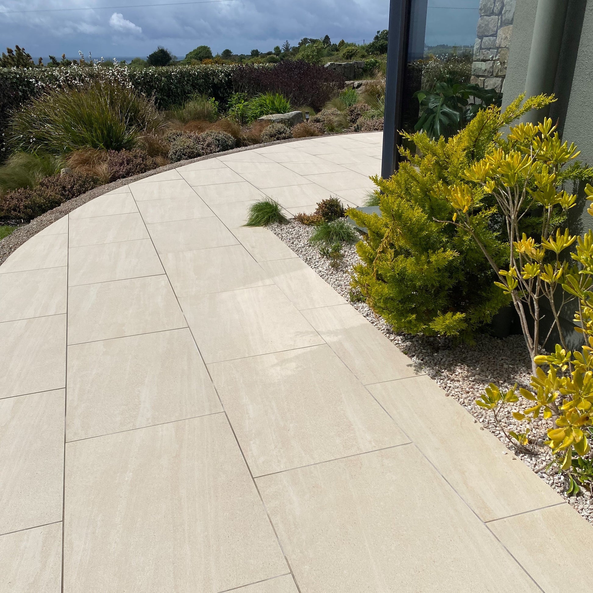 Paved pathway with garden and stone wall in the background. Patio tiles are Camel Jewel Range Porcelain Paving. 