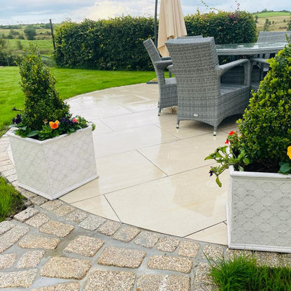 Outdoor patio area with wicker chairs, table, and potted plants in a garden setting. Paving is Camel Jewel Range Porcelain Paving Tiles with pathway and paving border completed in Gold Granite Cobbles. 