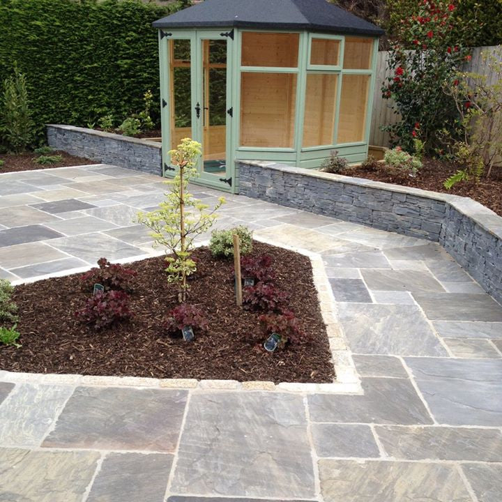 Paved patio area finished using Cider Black Sandstone patio packs surrounded by a small garden and wooden outdoor room in the background. 