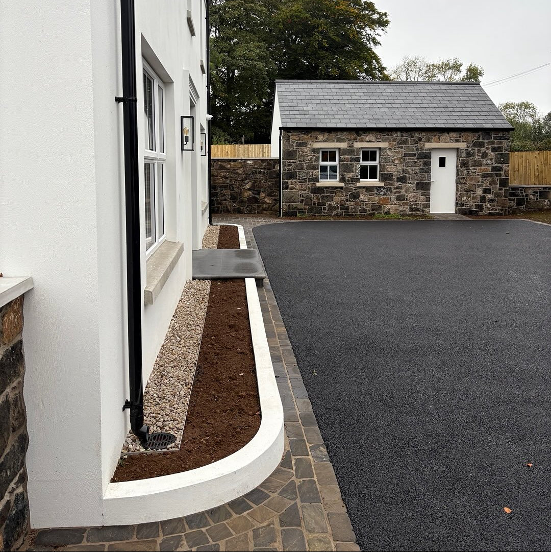 White house exterior with a tarmac driveway leading to a stone building. Tarmac has a cobbled border featuring Cider Black Sandstone Paving Setts. 