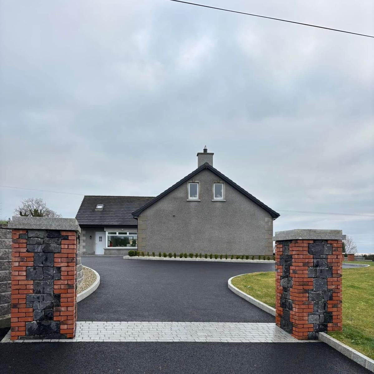 Tarmac driveway leading to a house with brick pillars on a cloudy day. Entrance features Coachman's Silver Granite Paving Setts. 