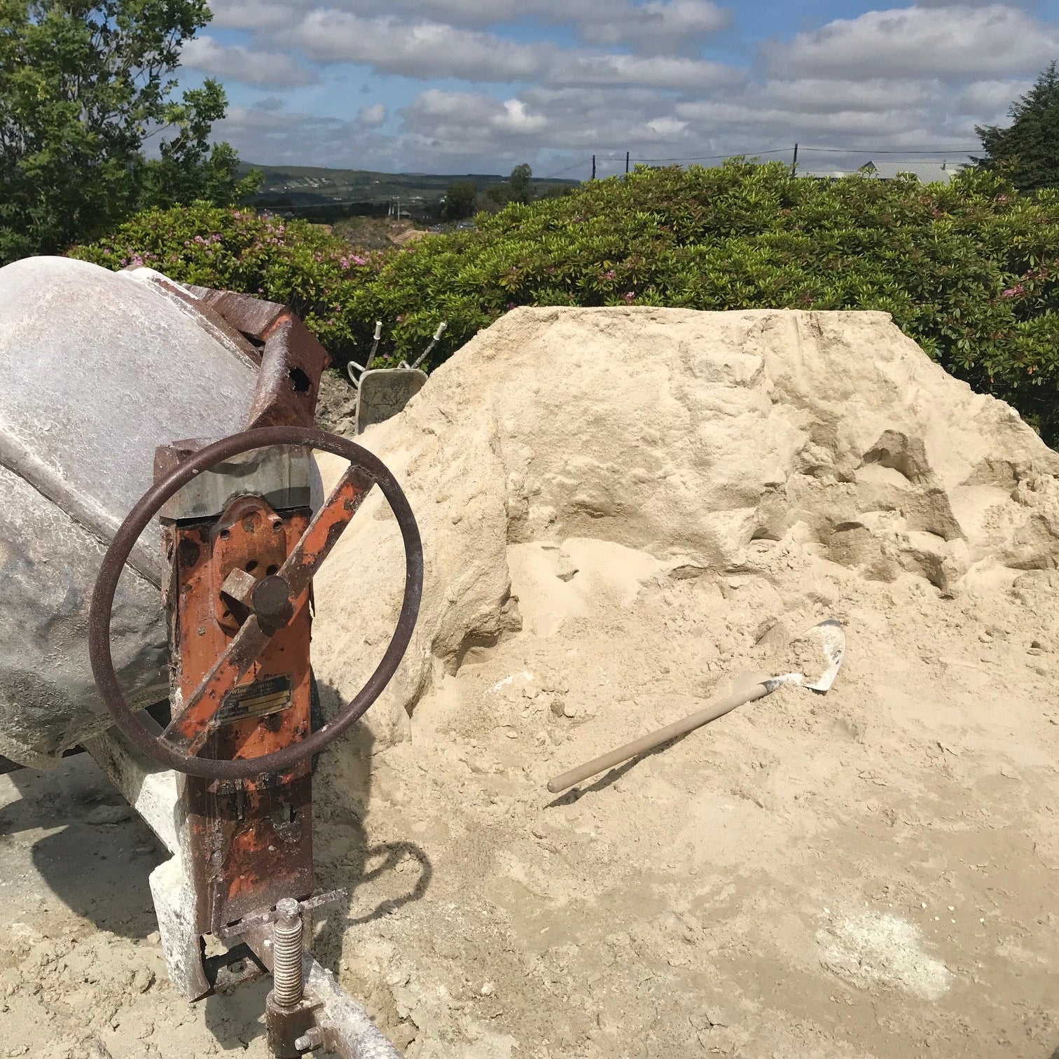 Rust-covered concrete mixer on a sandy surface with greenery in the background