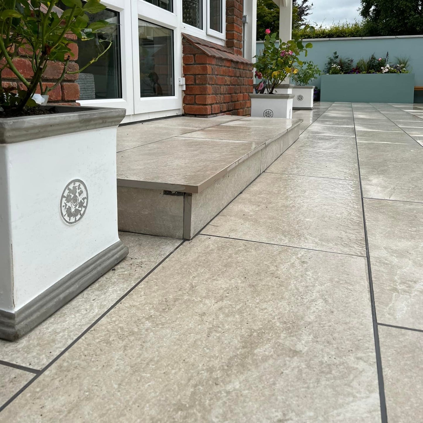 Close up of Cream Twice Qz Range Porcelain Paving Tiles installed on patio and step of a red brick house with potted pants and blue wall in the background. 