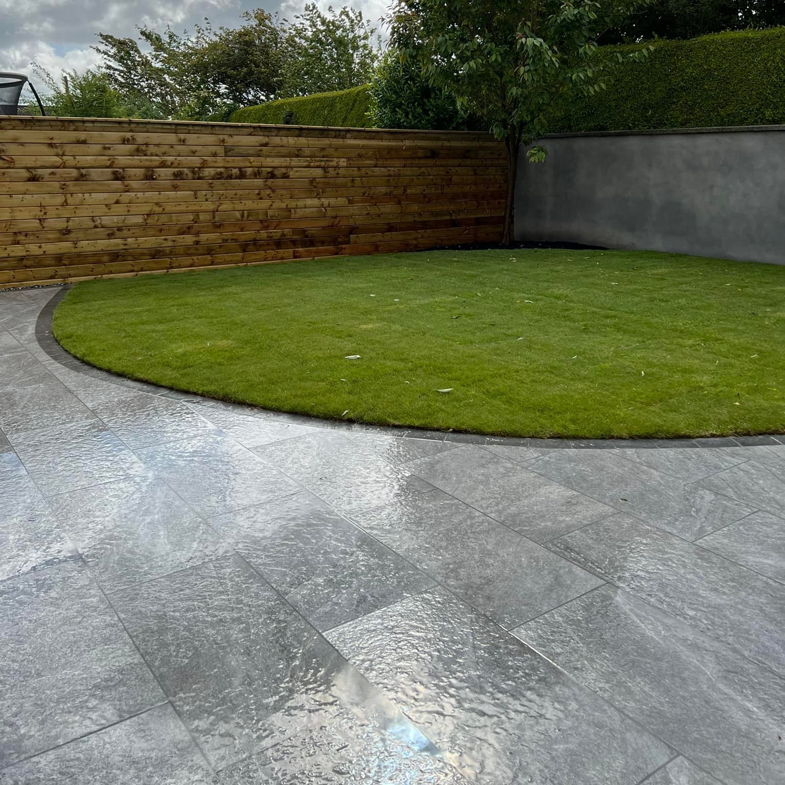 Patio with curved garden and large wooden fence and wall in the background. Paving is Dark Twice Qz Range Porcelain Paving Tiles. 
