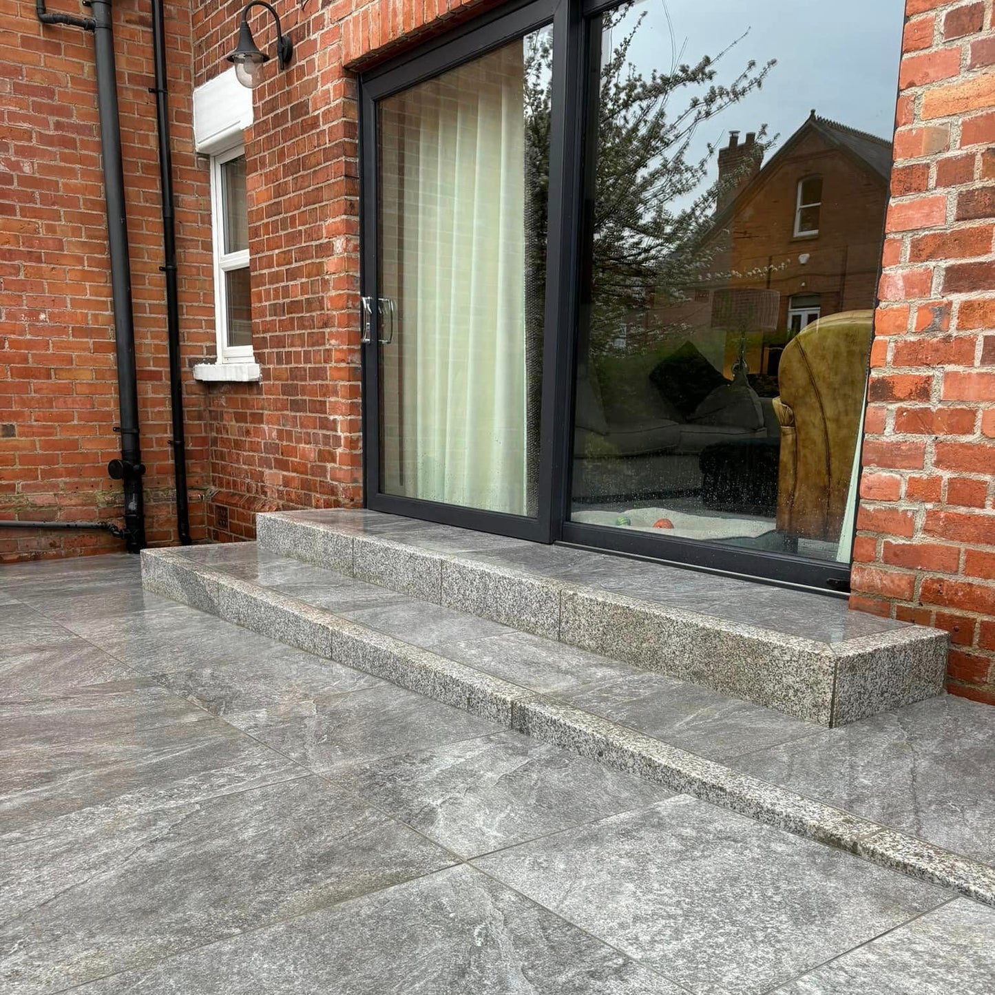 Red prick house with patio door leading out to steps and patio featuring Dark Twice Qz Range Porcelain Paving Tiles. 