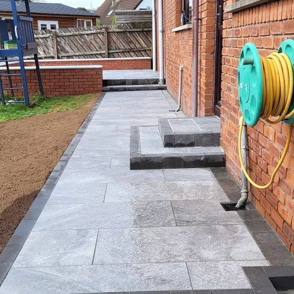 Pathway featuring Dark Twice Qz Range Porcelain Paving Tiles leading up to patio area of red brick house. 