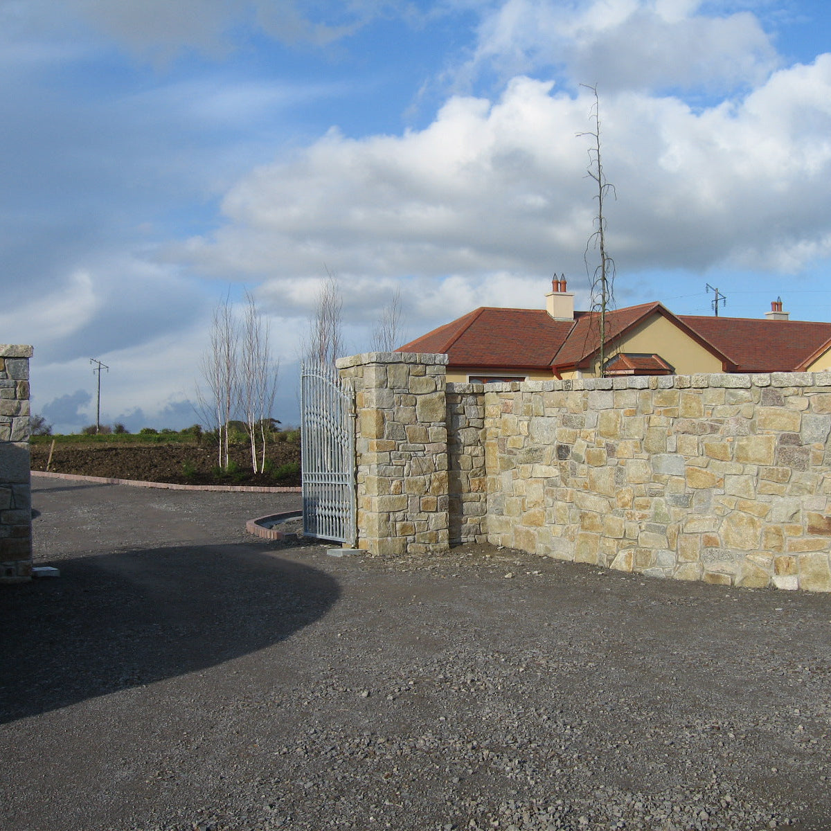 Stone wall in Donegal Granite with a gate leading to a residential area under a cloudy sky.