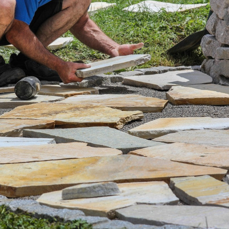 A person is laying stone down on a garden pathway, arranging Donegal Quartzite Crazy Paving to create a paving pattern. The stones are in various shades of brown and grey.