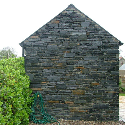 Stone-clad building in Donegal Slate Rubble with a green hose on a gravel surface, surrounded by greenery.
