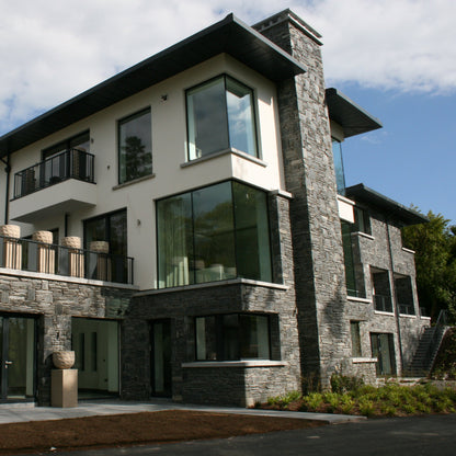 Modern house with stone façade in Donegal Slate Rubble surrounded by trees on a cloudy day.
