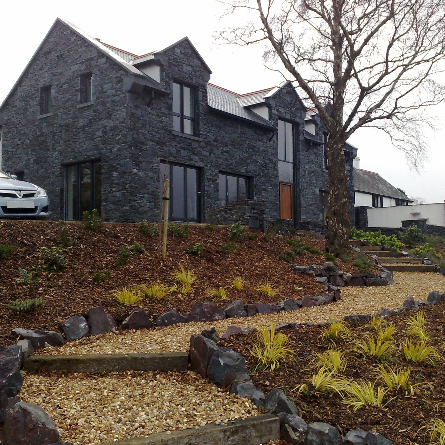 Modern stone house in Donegal Slate Rubble with landscaped garden featuring rocks and young trees.
