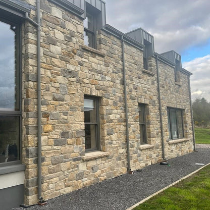 A exterior wall of a building featuring Dunhill Blend Sandstone with a tumbled finish, displaying golden brown, beige, and blue tones.