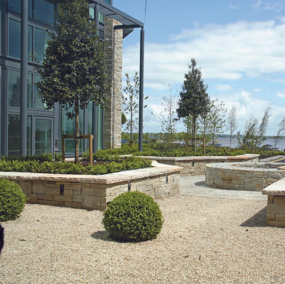 Outdoor area with low stone walls completed in Gold Donegal Quartzite Machined, greenery, and a building in the background. 