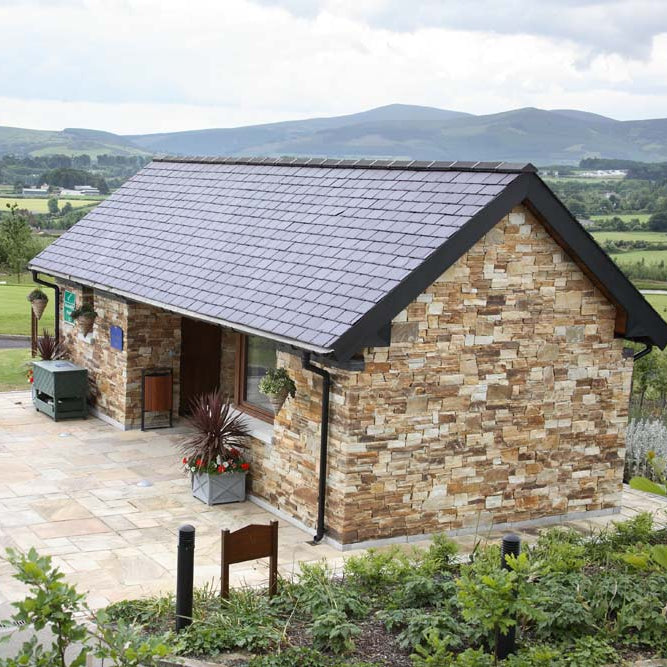 Modern stone building completed with Gold Donegal Quartzite Machined walling stone and a tiled roof in a scenic golf landscape with green fields and mountains.