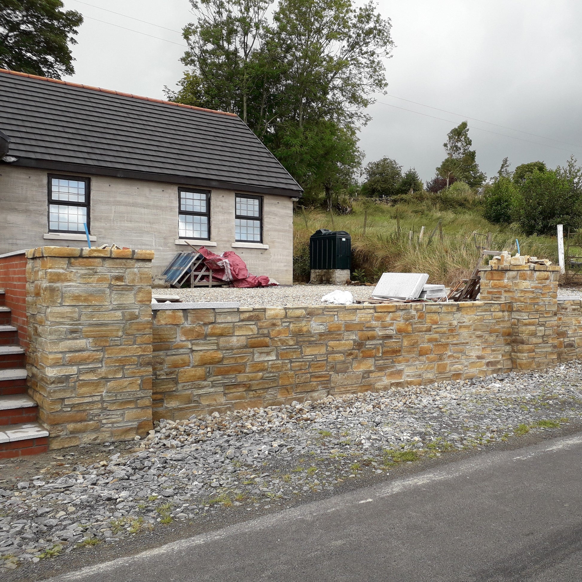Stone wall completed using Gold Donegal Quartzite Machined walling stone in front of a house with trees and a cloudy sky in the background.