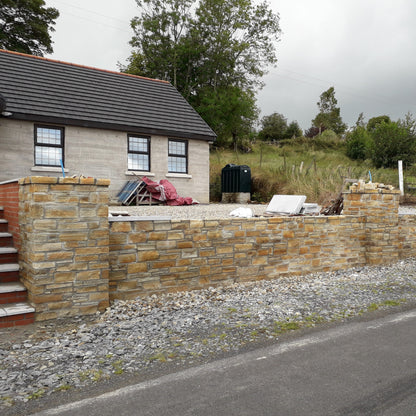 Stone wall completed using Gold Donegal Quartzite Machined walling stone in front of a house with trees and a cloudy sky in the background.