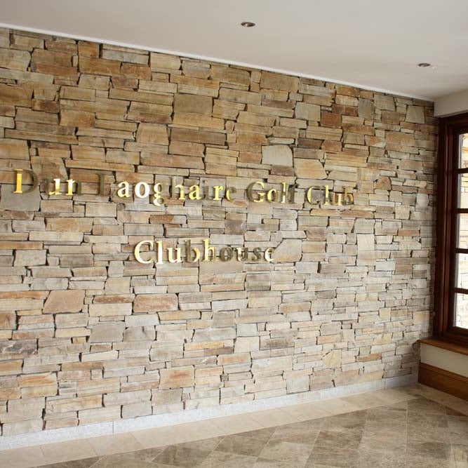Stone wall completed in Gold Donegal Quartzite Machined walling stone with 'Dun Laoghaire Golf Club Clubhouse' text in a well-lit room with large windows.