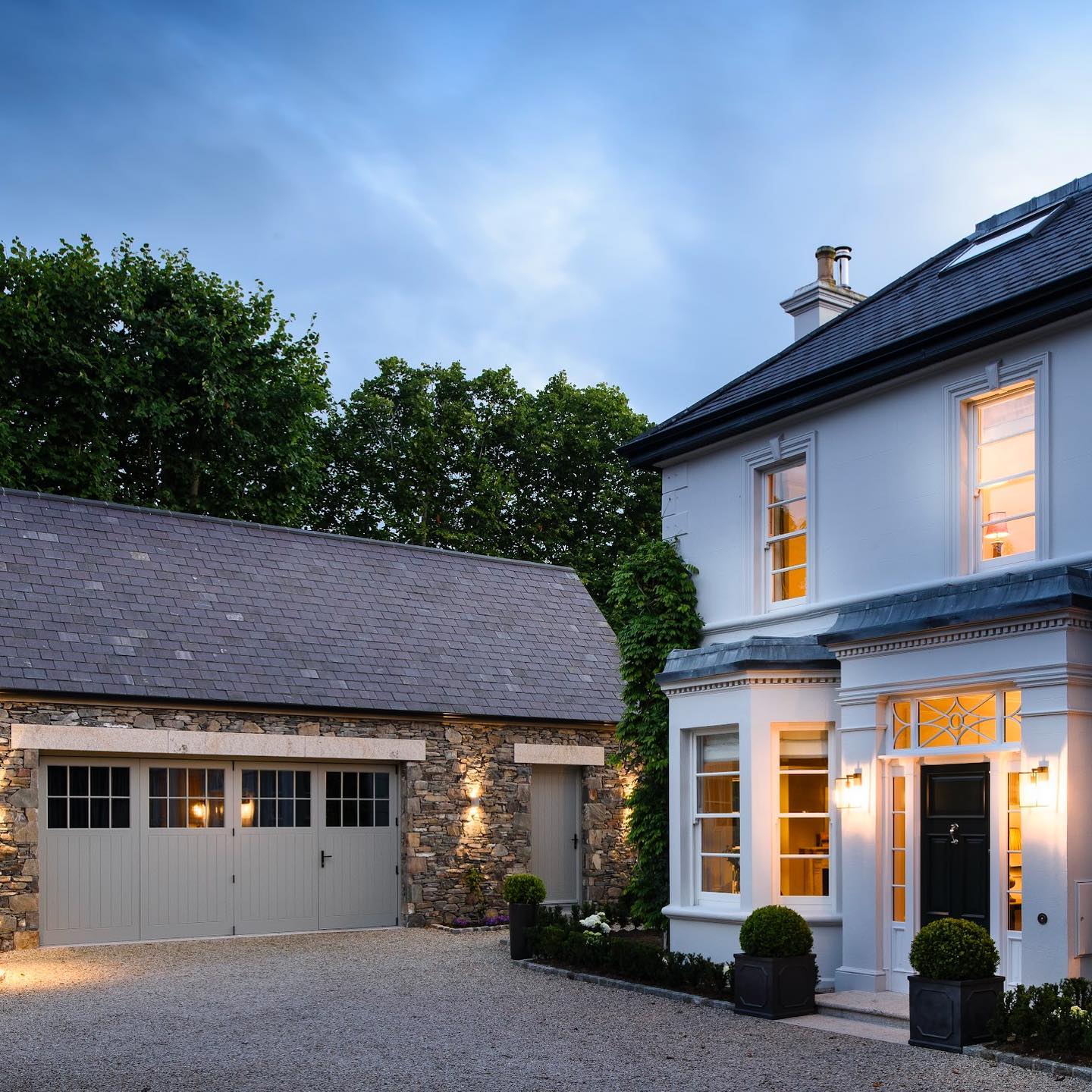 Stately home with a stone garage and illuminated windows on a clear evening. Stone garage featuring bespoke gold granite lintels. 