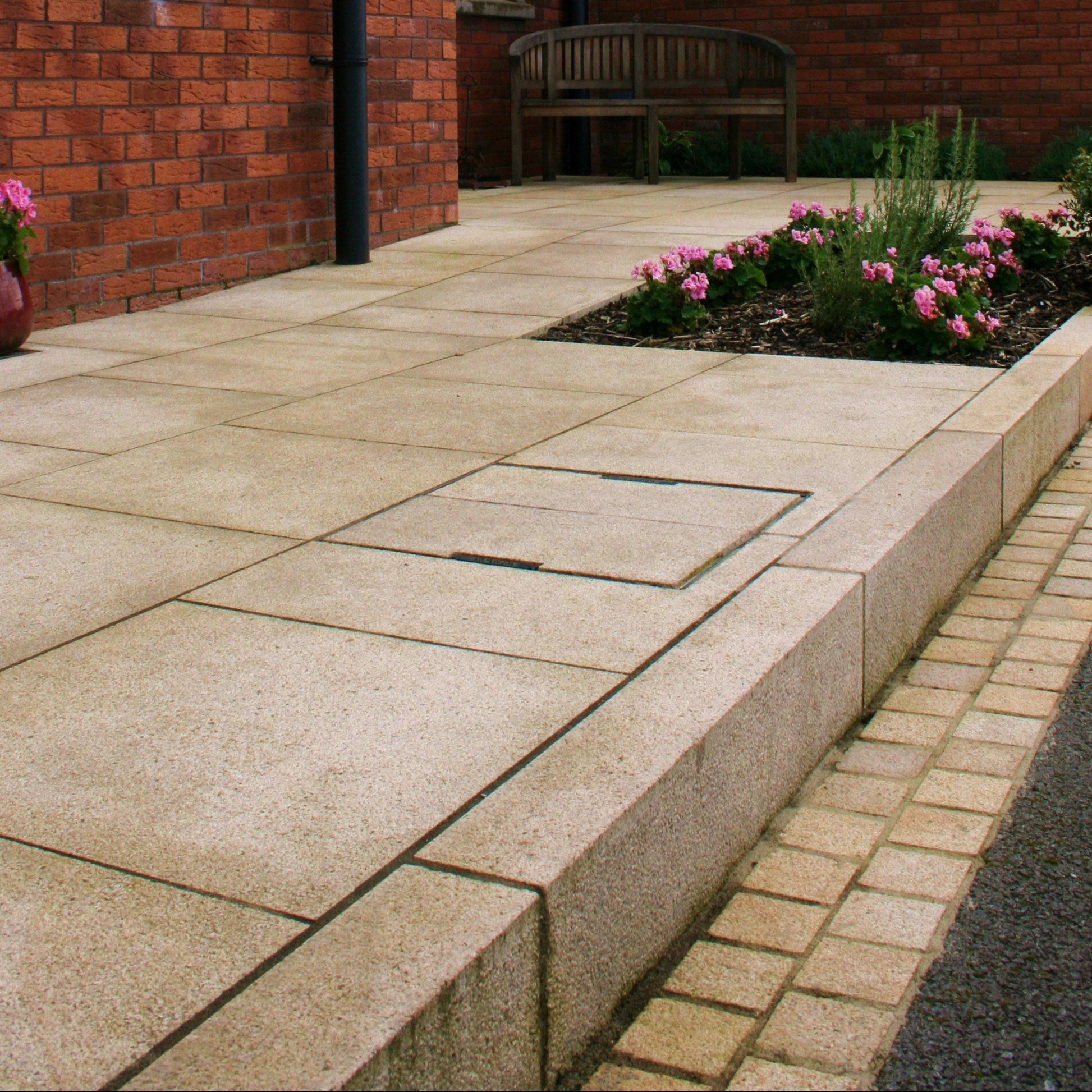Red brick house with gold granite paving and kerbing leading down to driveway. Bench and flowers in the background. 
