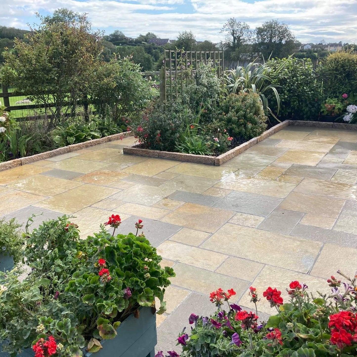 Paved patio area with garden plants and flowers, surrounded by greenery and Gold Granite Kerbs with a pineapple finish. 