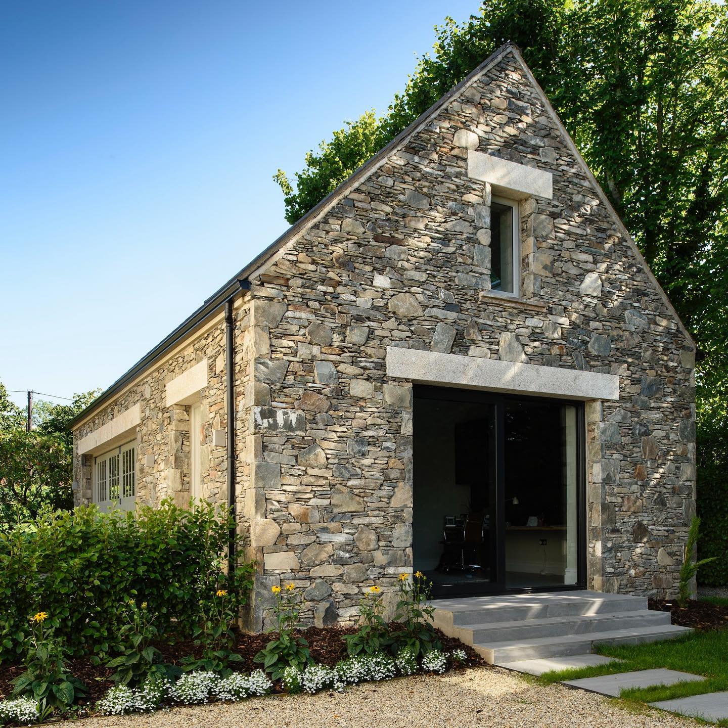 Stone building with a sloped roof and glass door surrounded by greenery. Stonework features gold granite lintels. 