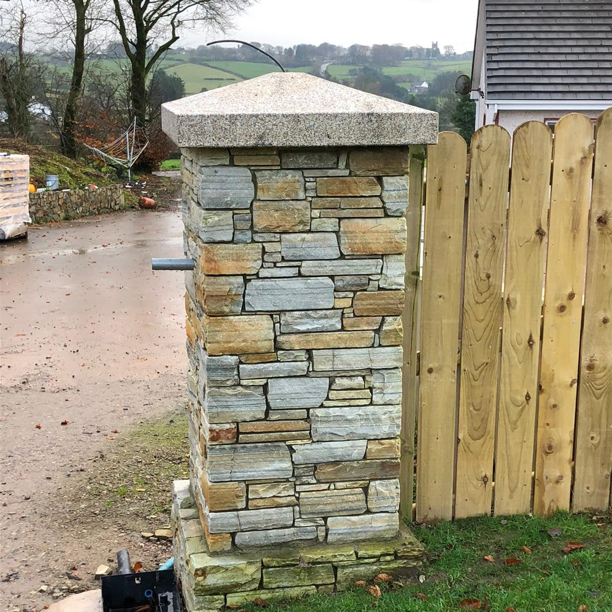 Stone pillar in a garden setting with a wooden fence and grass. Pillar features bespoke Gold Granite Apex Cap. 