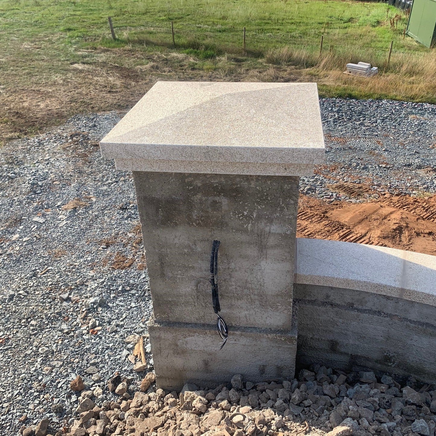 Bespoke Gold Granite Pillar and Wall Capping newly installed to concrete wall structure on a gravel surface with grass and a fence in the background. 