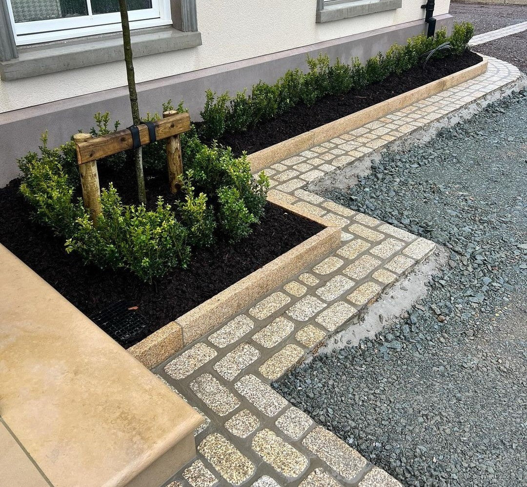 Paved pathway with landscaped garden area in front of a building featuring Gold Granite Pin Kerbs and Coachman's Cobbles. 