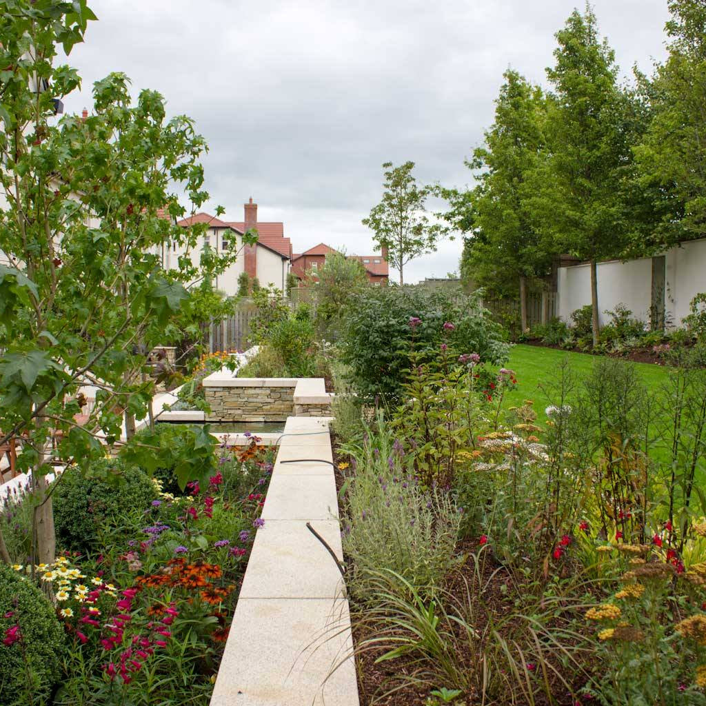 Garden surrounded by plants and flowers, with buildings in the background. Garden Walls feature Gold Granite Wall Capping. 