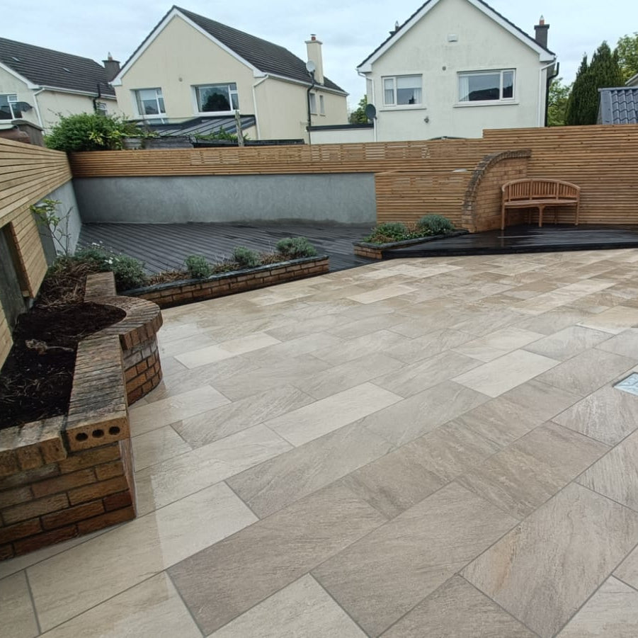 Backyard with stone patio, wooden deck, and garden features. Paving showcases Gold Quartz Diva Range Porcelain Paving. 