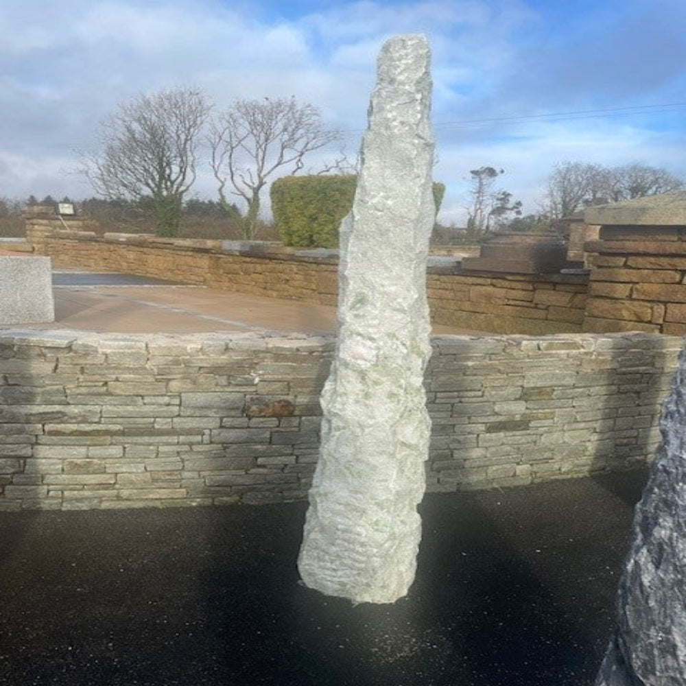Tall Green Marble Monolith in a landscaped area with a stone wall and trees in the background.