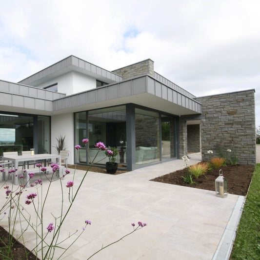Modern house with large glass windows and outdoor seating area, featuring flowers in the foreground. Stone is Grey Sandstone Machined. 