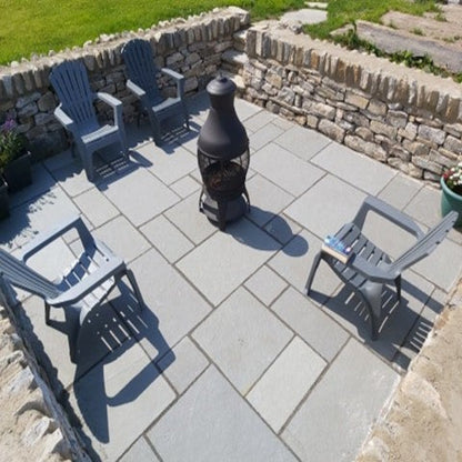 An outdoor patio area featuring grey sandstone paving slabs with a fire pit in the centre and four chairs placed on the slabs, set against a stone wall with a potted plants. 