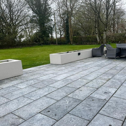 An outdoor paving area featuring Irish Blue Limestone slabs with a flamed finish, arranged on the ground in a patio setting with trees and a wall in the background.